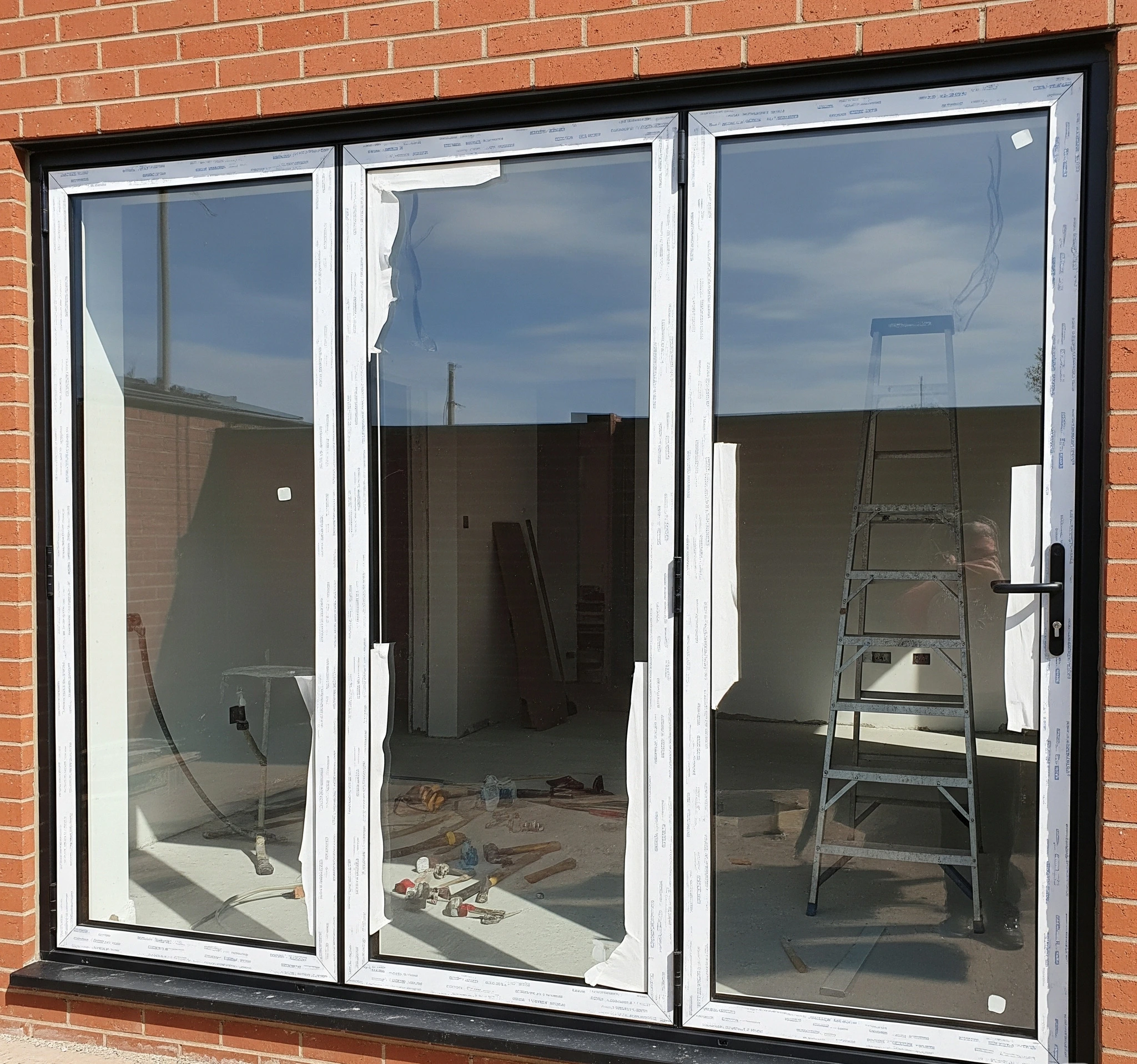 
Exterior view of a three-panel bi-fold door with white frames and a black outer trim, installed in a red brick wall, showing an unfinished interior.